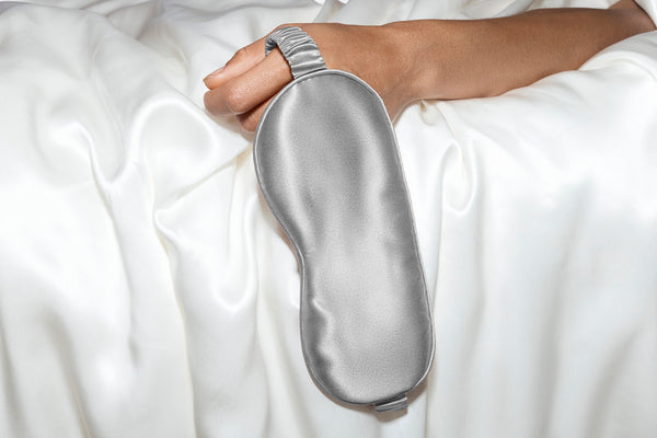 A hand holding a silver slip silk sleep mask on a white background.