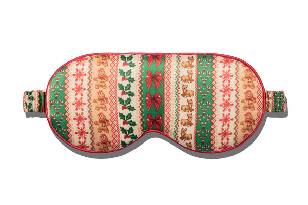 A festive Christmas-themed sleep mask with a pattern of gingerbread men, snowflakes, holly leaves, and red bows on a cream background, featuring red piping.