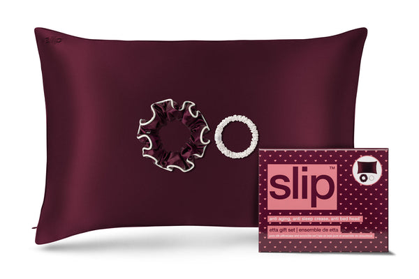 A crimson slip silk pillowcase with two scrunchies, shown next to its retail packaging box featuring the brand name 'slip'.
