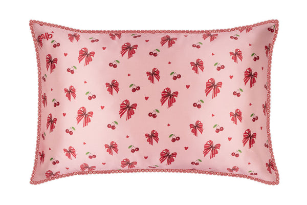 A slip silk pillowcase featuring a repeating pattern of red bows, cherries, and small hearts, finished with a scalloped lace trim.