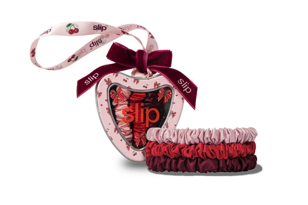 A heart-shaped Slip scrunchie gift set containing three silk scrunchies in pink, red, and burgundy, packaged in a heart-shaped tin box with a dark burgundy ribbon and a matching printed hanging ribbon.